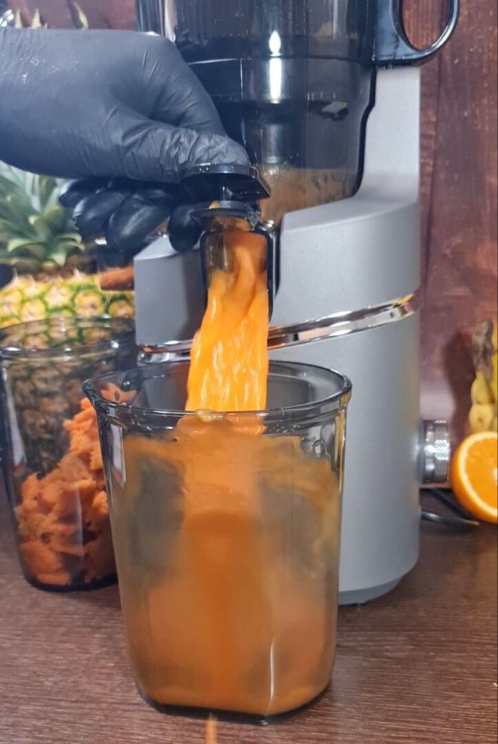 Fresh carrot juice being poured into a glass from an AMZCHEF juicer, surrounded by fresh ingredients including pineapple and oranges.