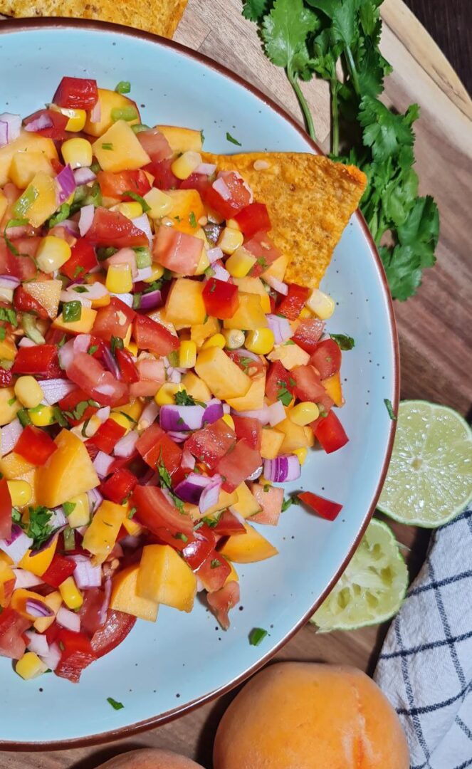 : A bowl of colorful peach salsa with tortilla chips, featuring tomatoes, peaches, corn, red onion, and cilantro.