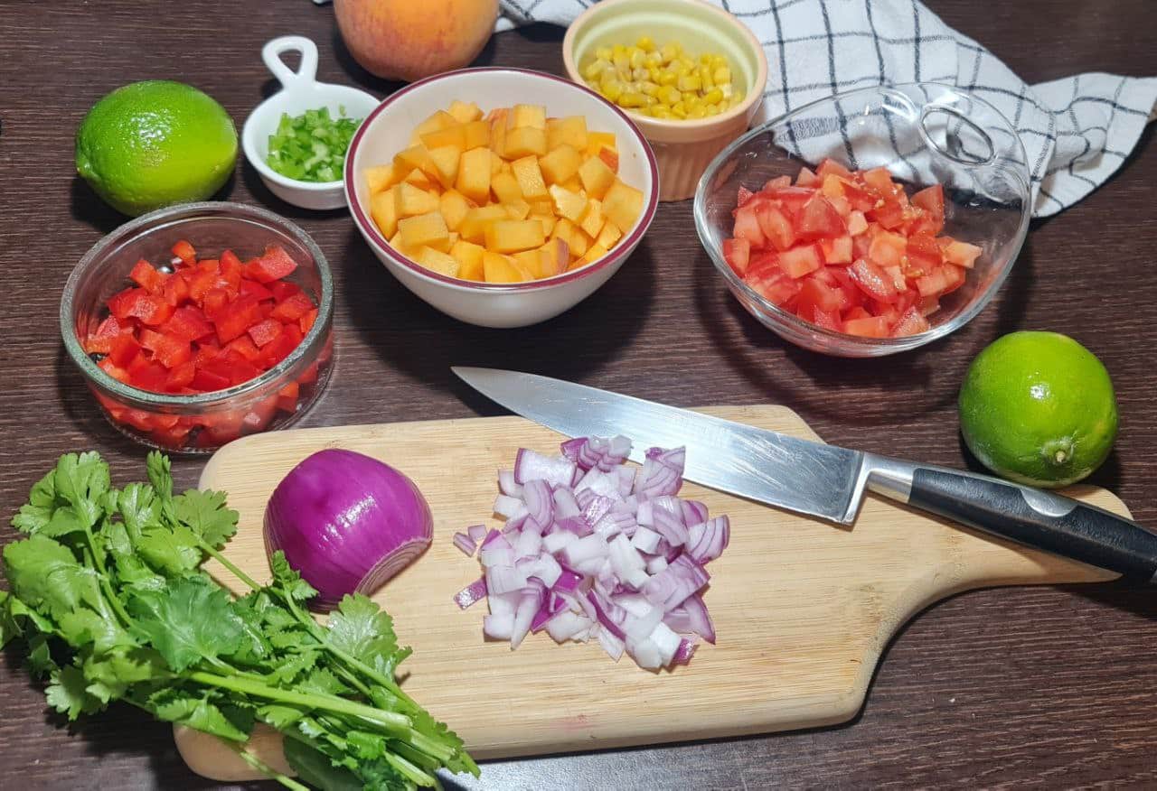  Chopped ingredients for peach salsa including red bell peppers, peaches, tomatoes, red onion, lime, cilantro, and corn.