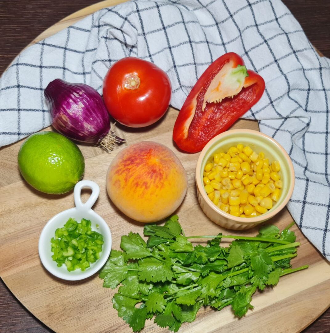  Whole ingredients for peach salsa on a wooden cutting board, including a peach, tomato, red bell pepper, red onion, lime, corn, jalapeno, and cilantro.