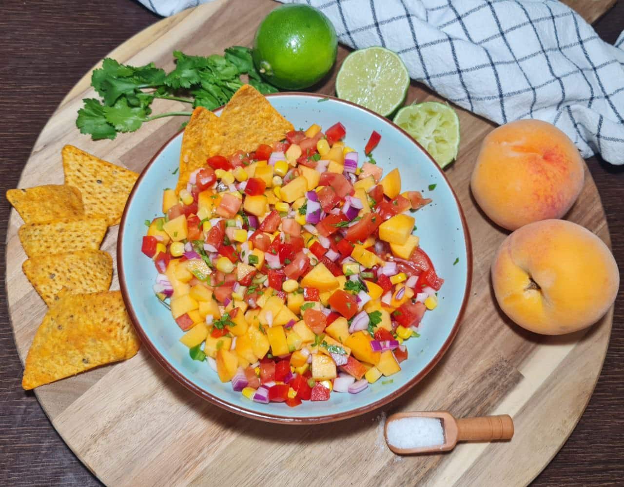 A bowl of peach salsa served with tortilla chips, surrounded by fresh peaches, lime, and cilantro.