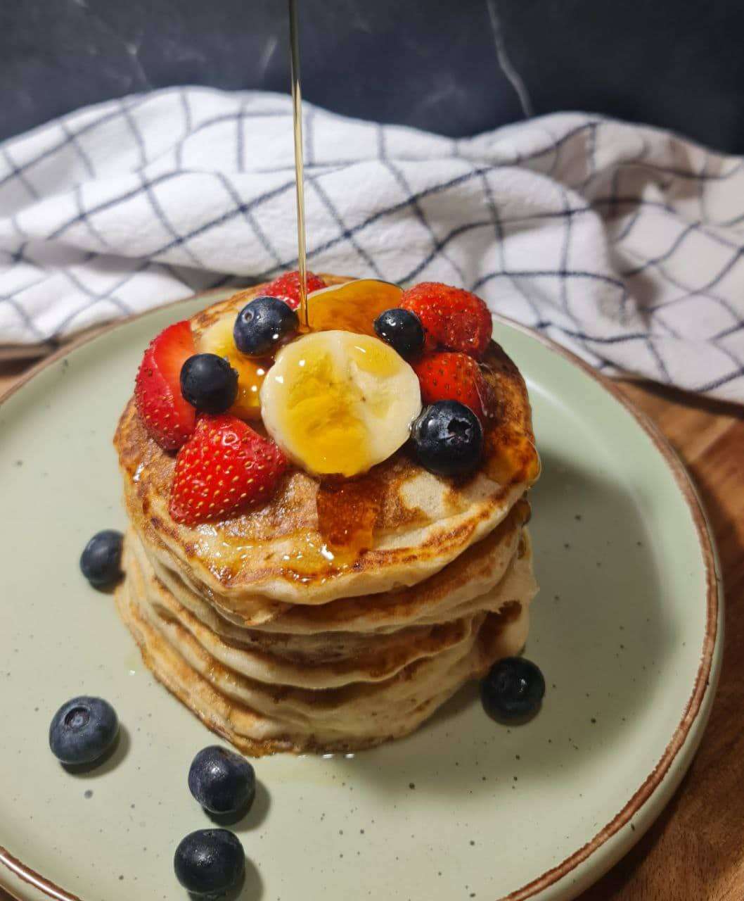 Stack of vegan banana pancakes topped with fresh strawberries, blueberries, banana slices, and a drizzle of syrup