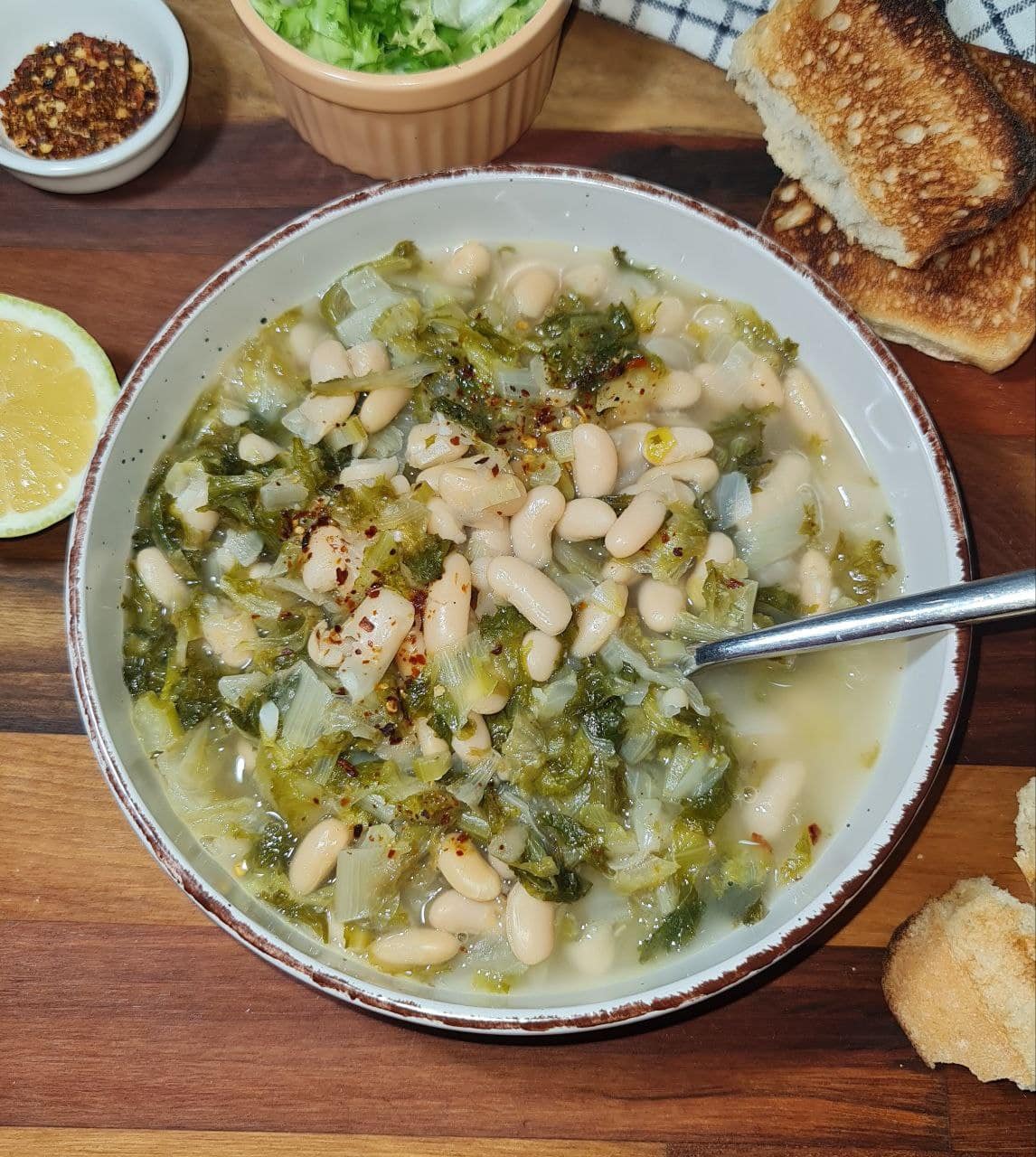 A bowl of Escarole White Bean Soup, garnished with red pepper flakes, served with toast and a side of lemon.