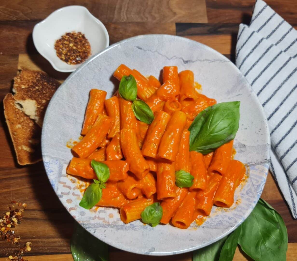 Creamy roasted red pepper pasta garnished with fresh basil leaves served in a white bowl, accompanied by toasted bread and red pepper flakes. A delicious and creamy roasted red pepper pasta, garnished with fresh basil, served with toasted bread and a sprinkle of red pepper flakes.