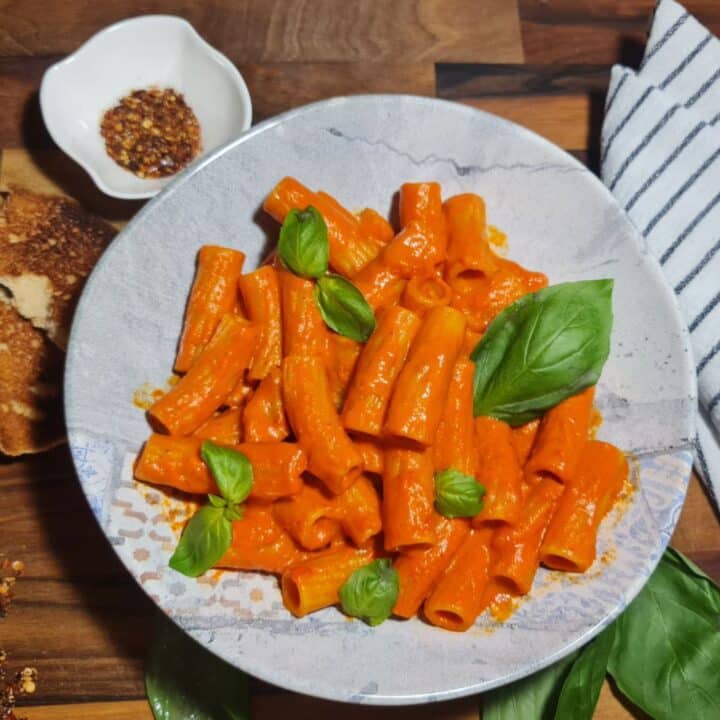 Creamy roasted red pepper pasta garnished with fresh basil leaves served in a white bowl, accompanied by toasted bread and red pepper flakes. A delicious and creamy roasted red pepper pasta, garnished with fresh basil, served with toasted bread and a sprinkle of red pepper flakes.