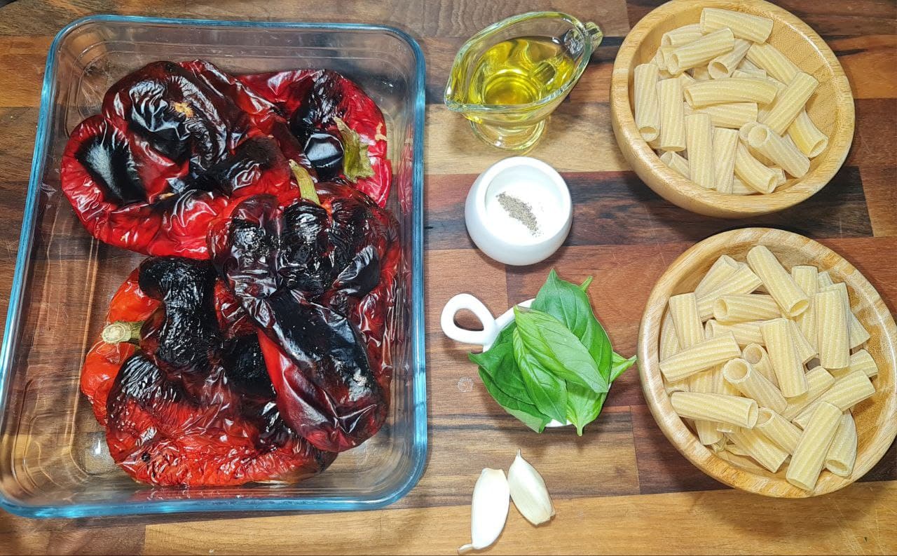 Ingredients for roasted red pepper pasta, including charred red peppers in a glass dish, olive oil, rigatoni pasta, fresh basil, garlic cloves, and a small bowl of salt and pepper. Key ingredients for roasted red pepper pasta: charred red peppers, rigatoni pasta, fresh basil, garlic, olive oil, and seasoning.