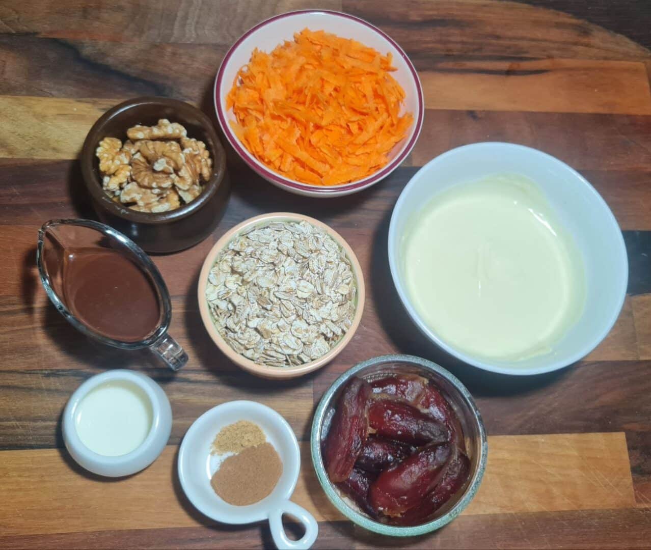 Ingredients for vegan carrot cake balls, including grated carrots, oats, dates, walnuts, white and dark chocolate, spices, and plant-based milk, arranged on a wooden surface.The wholesome ingredients for vegan carrot cake balls, featuring grated carrots, oats, dates, walnuts, white and dark chocolate, spices, and plant-based milk. 