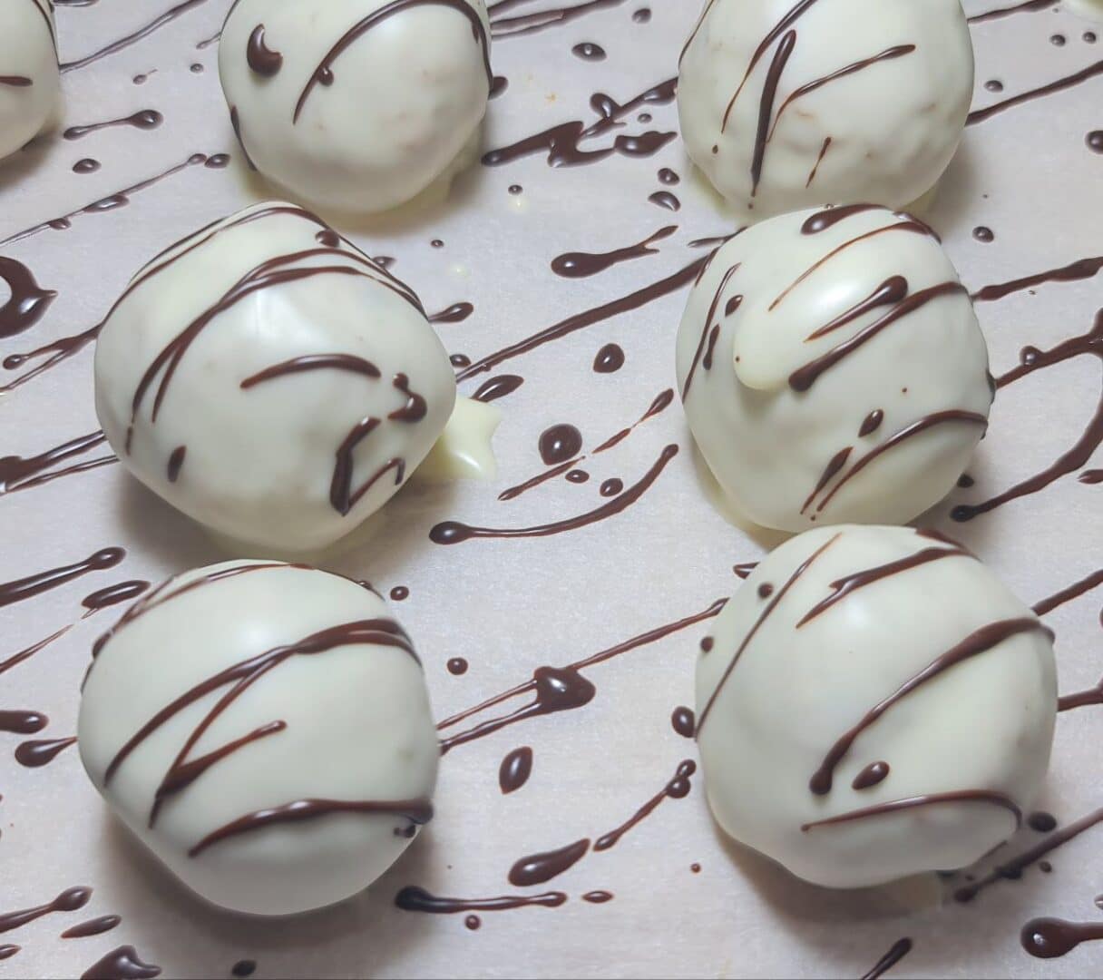  Vegan carrot cake balls coated in white chocolate with dark chocolate drizzle, arranged on parchment paper. Vegan carrot cake balls dipped in white chocolate, elegantly drizzled with dark chocolate, for a delightful treat.