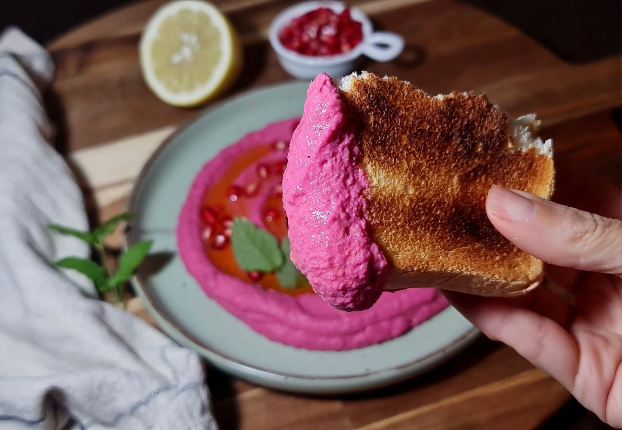 Crispy toast dipped in vibrant pink beetroot hummus. A slice of crispy toast dipped in creamy beetroot hummus, topped with pomegranate seeds.