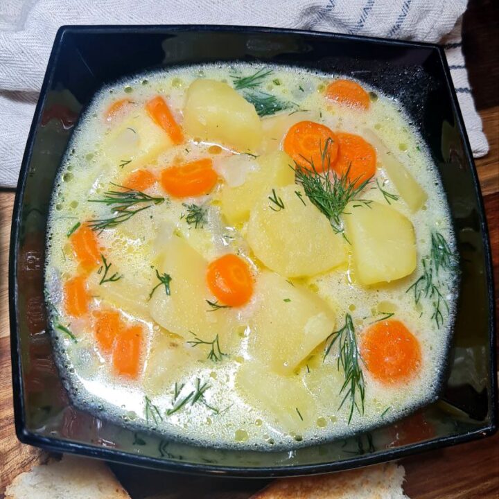 Creamy vegan potato dill soup with potatoes, carrots, and fresh dill served in a black bowl with toasted bread. A bowl of creamy vegan potato dill soup served with fresh dill and toasted bread on the side.