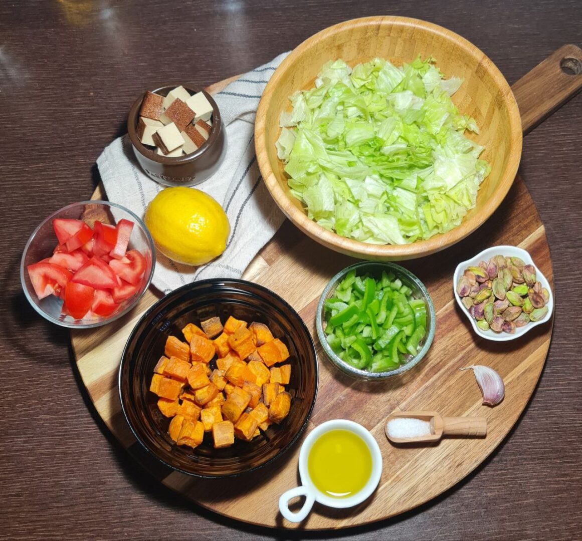 Ingredients for smoked tofu sweet potato salad including lettuce, diced tomatoes, green bell peppers, sweet potato cubes, smoked tofu, pistachios, olive oil, and a lemon arranged on a wooden cutting board.
Preparing the ingredients for a smoked tofu and sweet potato salad with fresh vegetables and nuts.