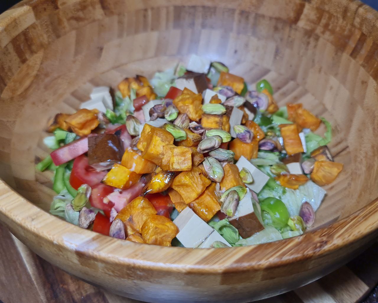 A wooden bowl filled with smoked tofu sweet potato salad with lettuce, tomatoes, roasted sweet potato cubes, pistachios, and smoked tofu. A vibrant smoked tofu and sweet potato salad with fresh vegetables and crunchy pistachios.