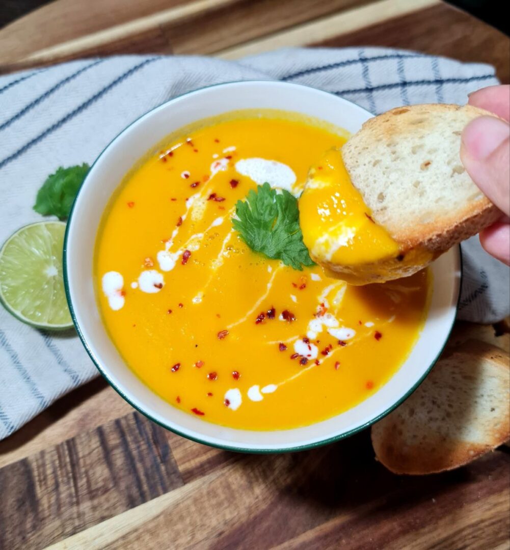 Hand dipping bread into a bowl of Thai coconut pumpkin soup garnished with cilantro, coconut cream, and chili flakes. Creamy Thai coconut pumpkin soup, perfect for dipping with crispy bread.