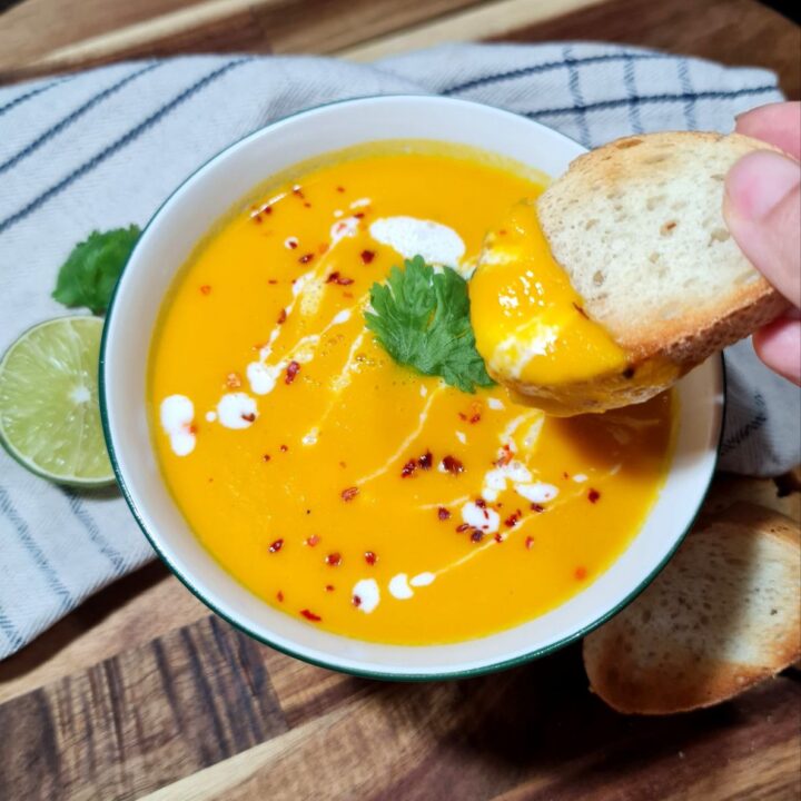 Hand dipping bread into a bowl of Thai coconut pumpkin soup garnished with cilantro, coconut cream, and chili flakes. Creamy Thai coconut pumpkin soup, perfect for dipping with crispy bread.