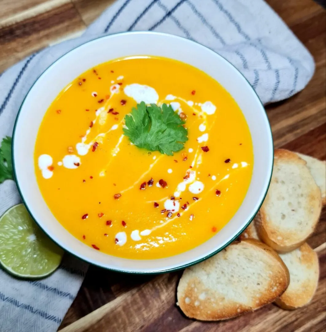 Bowl of Thai coconut pumpkin soup garnished with cilantro, coconut cream, and chili flakes, served with lime and toasted bread. Thai coconut pumpkin soup served with lime and toasted bread for a perfect fall meal.
