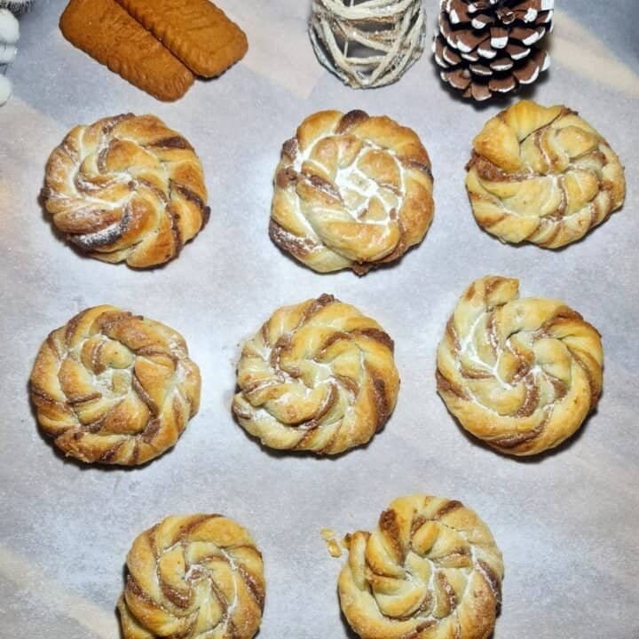 Eight round Biscoff swirls arranged on a parchment-lined baking tray with three rectangular cookies and decorative pine cones.