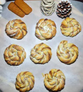 Eight round Biscoff swirls arranged on a parchment-lined baking tray with three rectangular cookies and decorative pine cones.