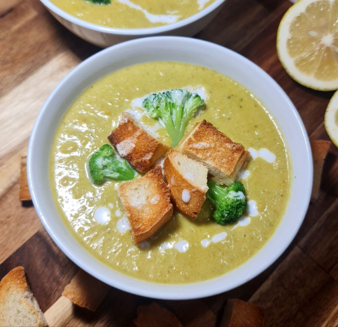 vegan broccoli cheddar soup, garnished with croutons and broccoli, placed on a wooden board with lemon slices. Delicious vegan broccoli cheddar soup, topped with croutons and broccoli florets, served on a wooden board with lemon slices.