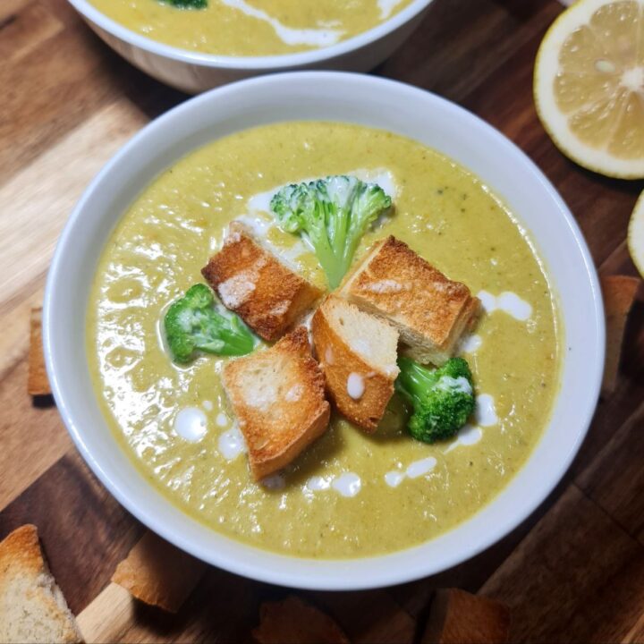 vegan broccoli cheddar soup, garnished with croutons and broccoli, placed on a wooden board with lemon slices. Delicious vegan broccoli cheddar soup, topped with croutons and broccoli florets, served on a wooden board with lemon slices.