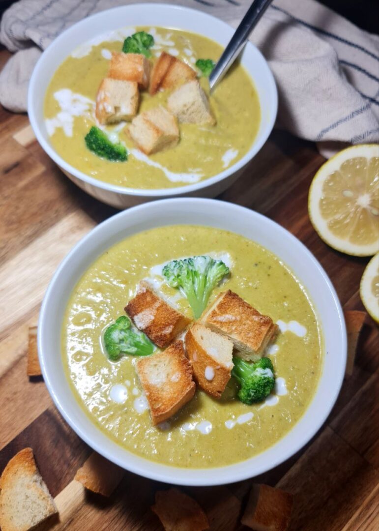 Vegan broccoli cheddar soup topped with croutons and broccoli florets, served in a white bowl with lemons on the side. A comforting bowl of vegan broccoli cheddar soup garnished with croutons and broccoli, with fresh lemons in the background.