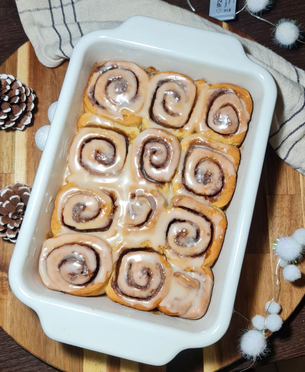 Freshly baked vegan cinnamon rolls with icing in a white baking dish, surrounded by pinecones and festive decorations.