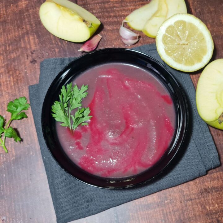 Vegan red cabbage soup garnished with parsley in a black bowl on a wooden table, surrounded by apple slices and garlic. A comforting bowl of vegan red cabbage soup with a touch of parsley for garnish.