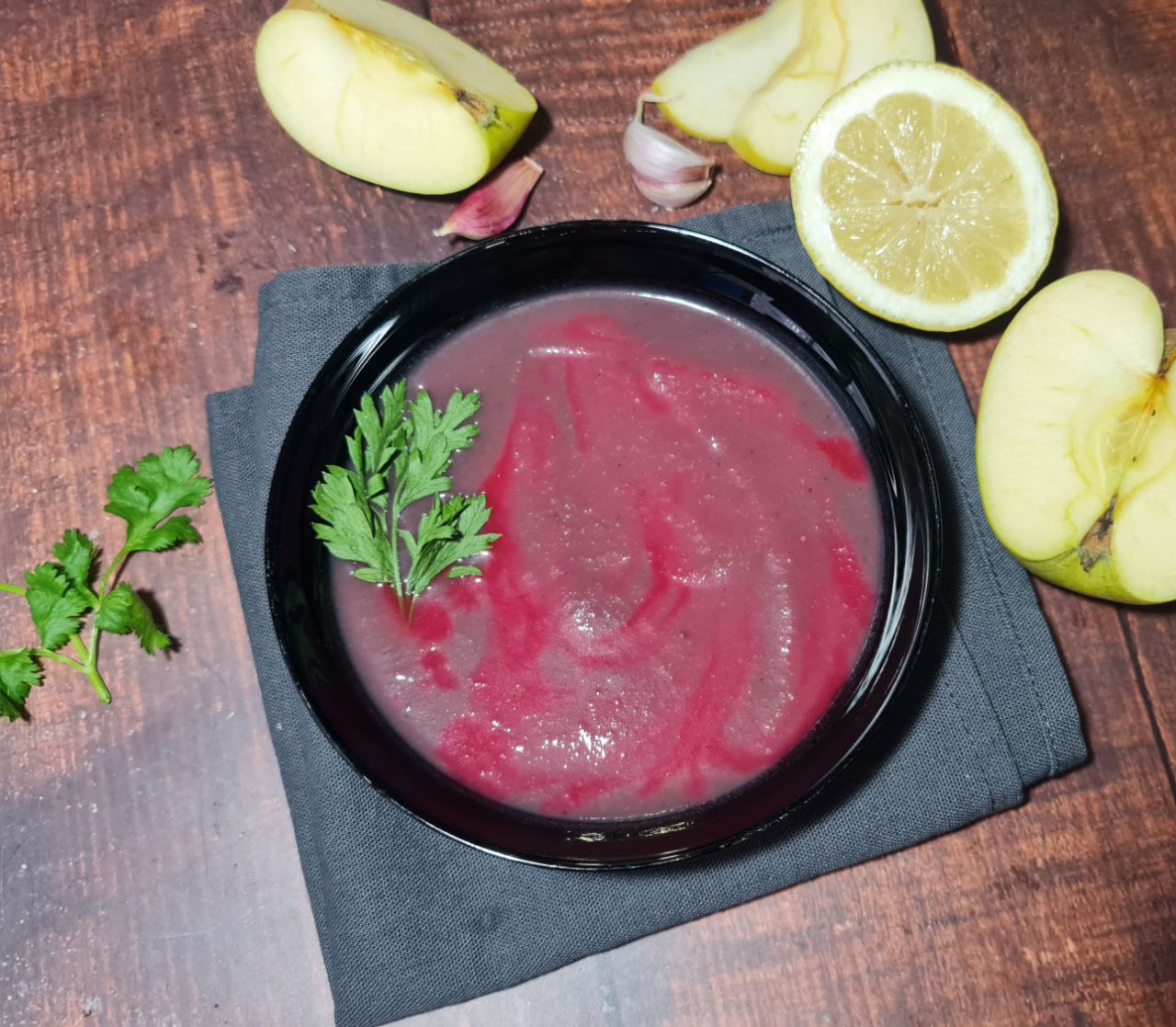 Vegan red cabbage soup garnished with parsley in a black bowl on a wooden table, surrounded by apple slices and garlic. A comforting bowl of vegan red cabbage soup with a touch of parsley for garnish.