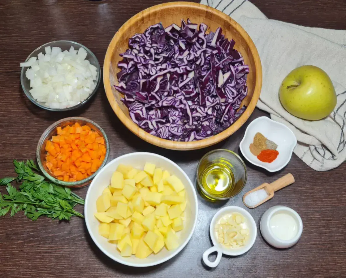 Ingredients for vegan red cabbage soup including red cabbage, potatoes, apples, carrots, onions, garlic, and spices laid out on a countertop.  The fresh ingredients needed for making vegan red cabbage soup, including red cabbage, potatoes, and spices.