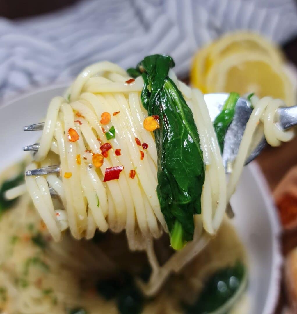 Close-up of a fork holding vegan spaghetti aglio e olio with fresh spinach and red pepper flakes. : A fork twirling vegan spaghetti aglio e olio with spinach, garnished with red pepper flakes.