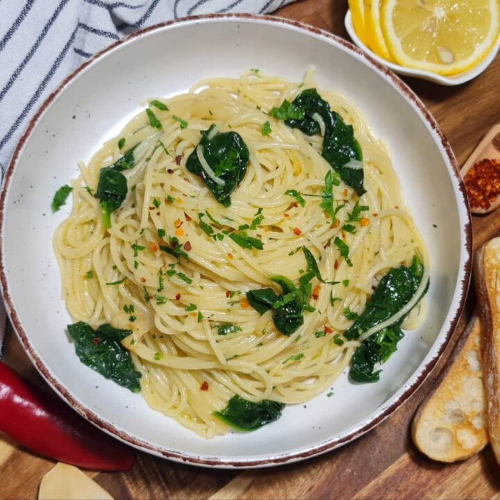 Top view of a plate of vegan spaghetti aglio e olio with spinach served with lemon slices, garlic, and bread. A serving of vegan spaghetti aglio e olio with spinach, complemented by lemon, garlic, and crispy bread on the side