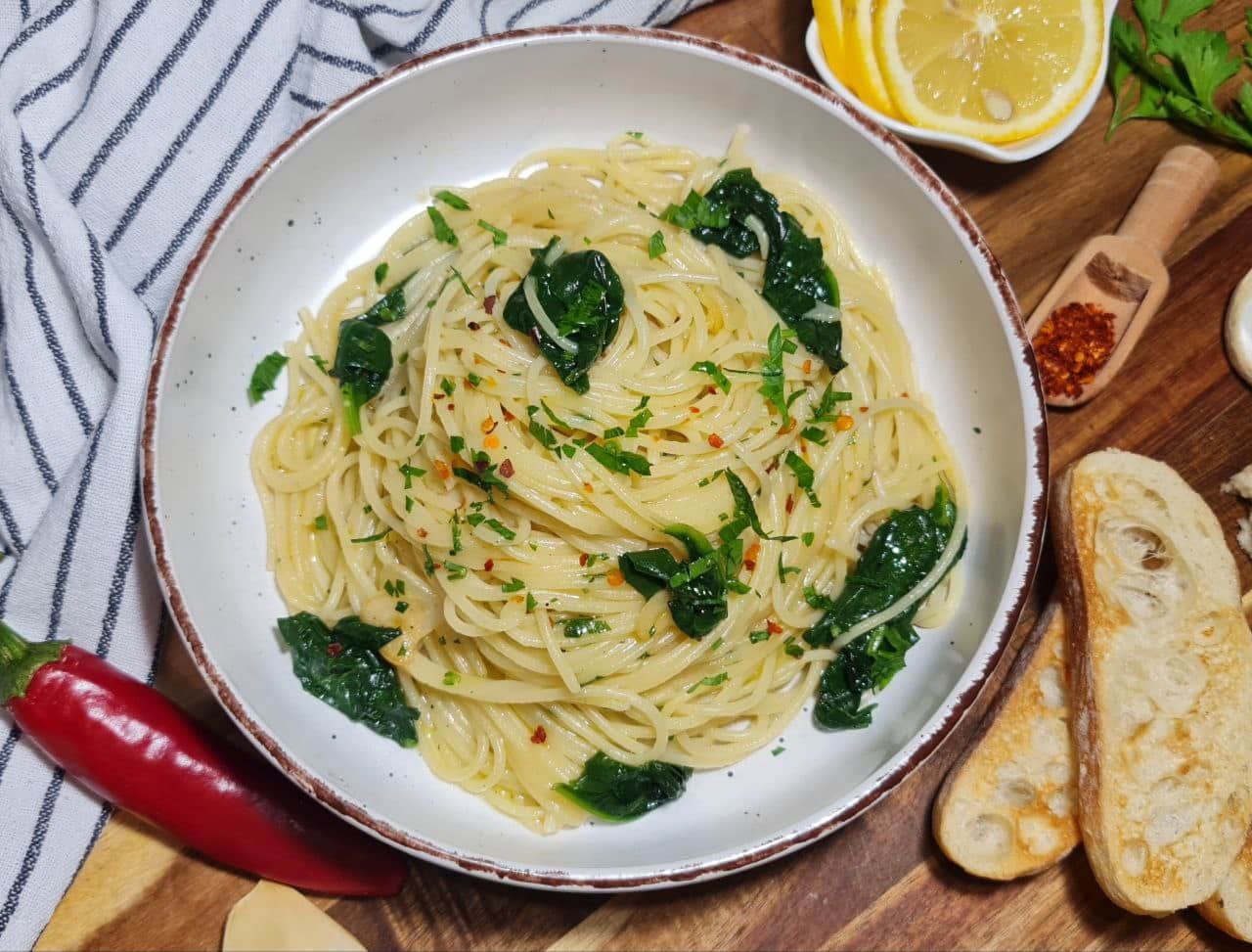 Top view of a plate of vegan spaghetti aglio e olio with spinach served with lemon slices, garlic, and bread. A serving of vegan spaghetti aglio e olio with spinach, complemented by lemon, garlic, and crispy bread on the side