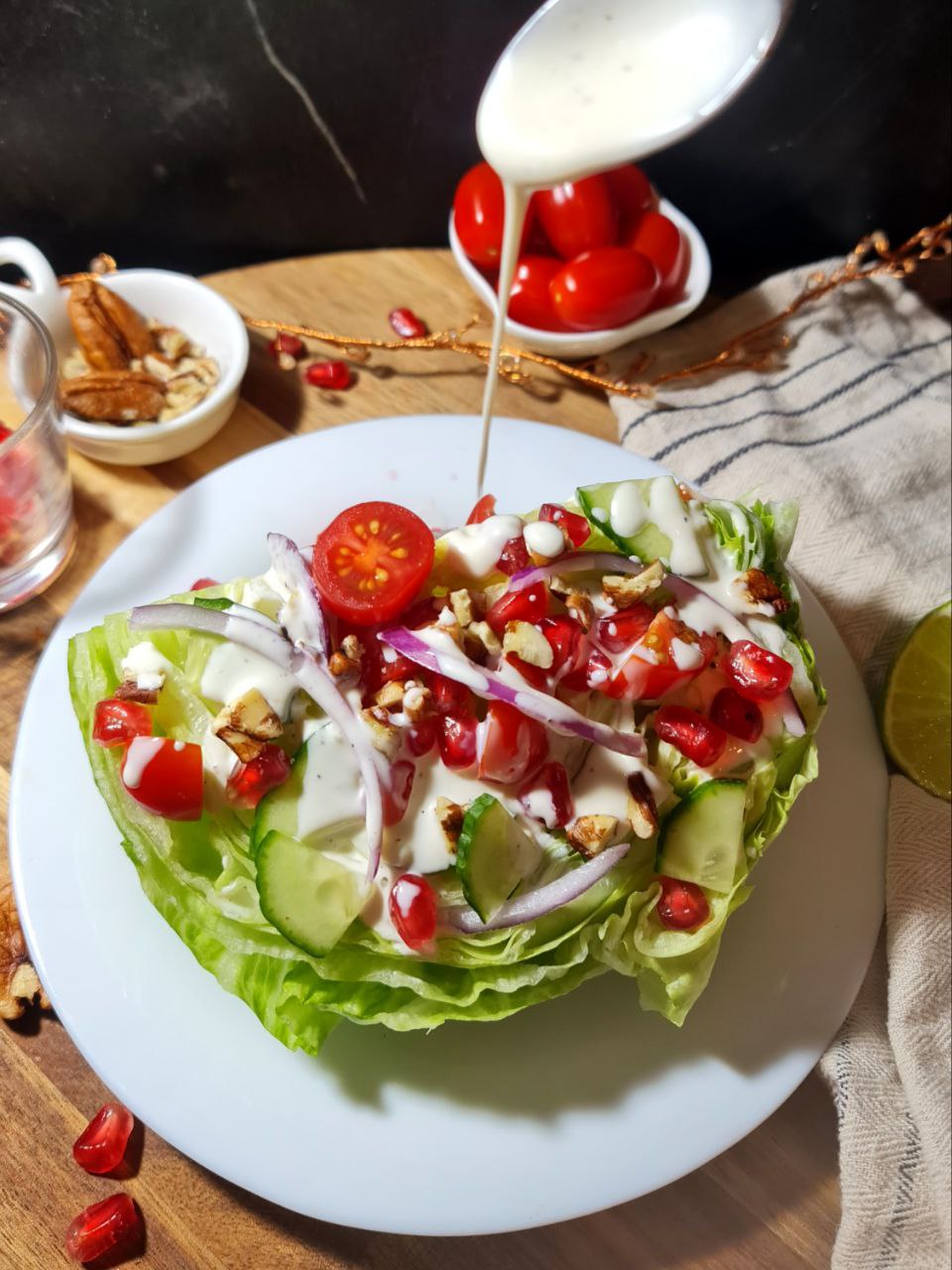 A vibrant vegan wedge salad topped with cherry tomatoes, red onions, cucumber, pomegranate seeds, and nuts, being drizzled with a creamy dressing.