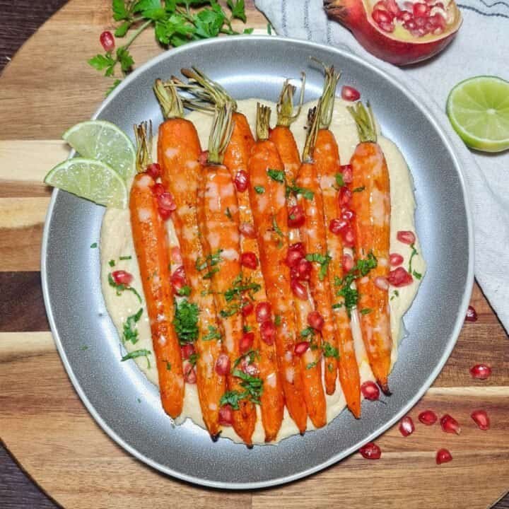 Roasted carrots served on a bed of creamy hummus, garnished with pomegranate seeds and fresh parsley. Whole roasted carrots with hummus, pomegranate, and parsley.