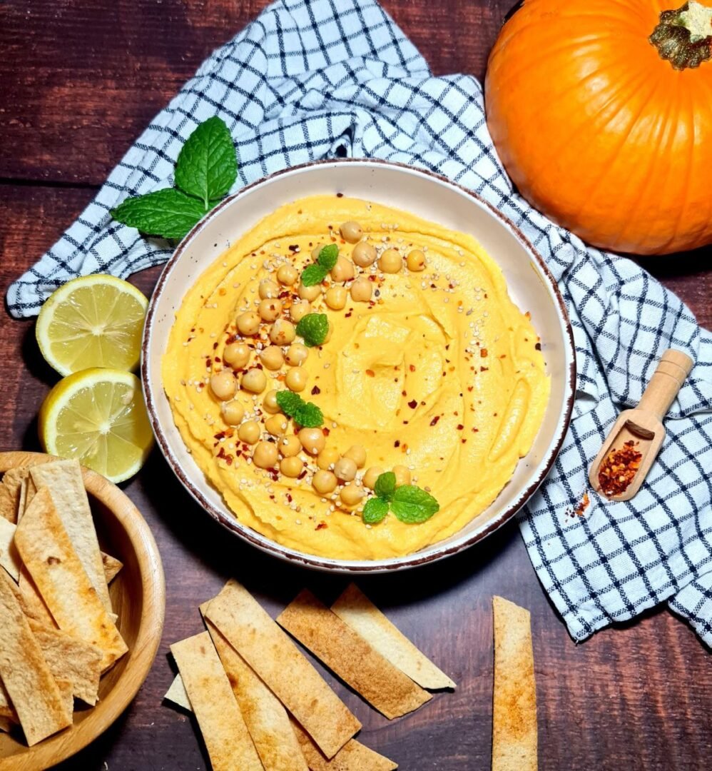 Bowl of pumpkin hummus garnished with chickpeas, mint, and red pepper flakes, served with lemon slices and tortilla strips. Enjoy this flavorful pumpkin hummus with crispy tortilla strips.