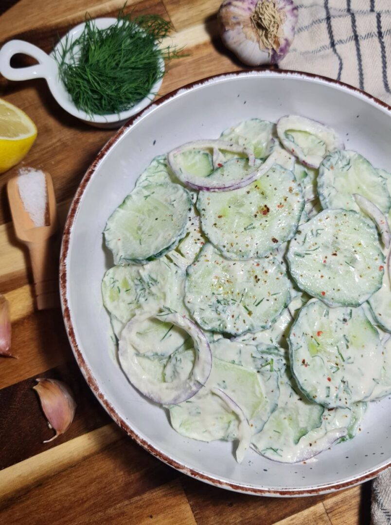 Creamy cucumber salad with red onions, fresh dill, and a tangy dressing in a white bowl. Finished creamy cucumber salad topped with red onions and fresh dill.