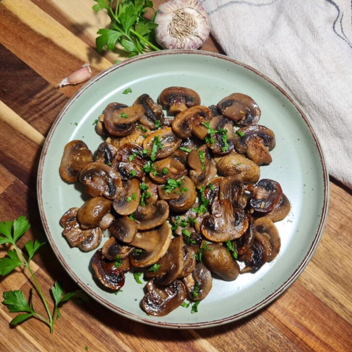 Plate of sautéed mushrooms garnished with parsley on a wooden board. A plate full of sautéed mushrooms, garnished with parsley, served as a savory vegan side dish.