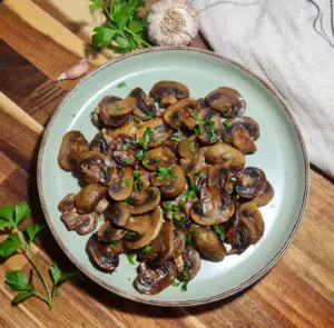 Plate of sautéed mushrooms garnished with parsley on a wooden board. A plate full of sautéed mushrooms, garnished with parsley, served as a savory vegan side dish.