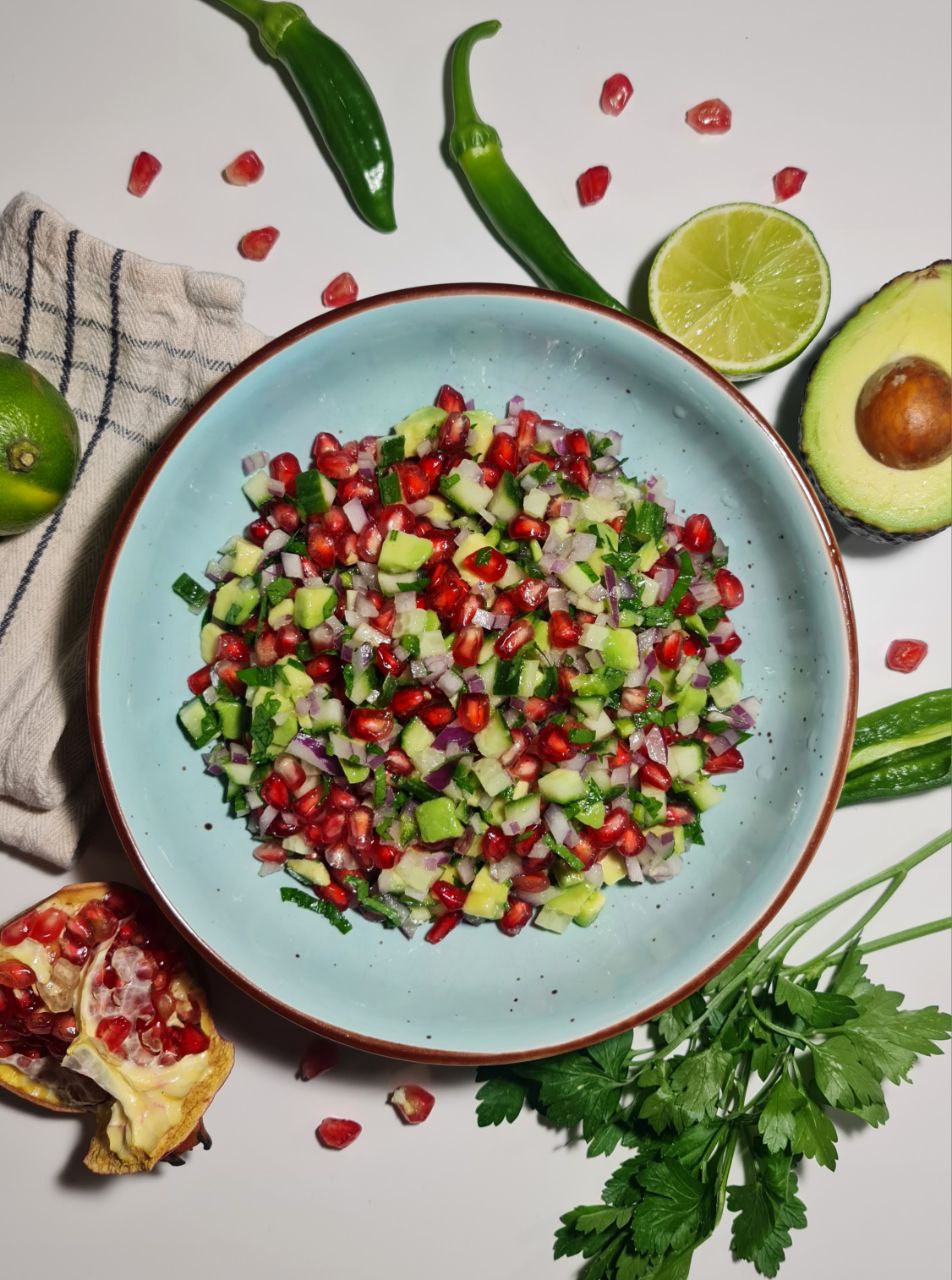 A bowl of colorful pomegranate salsa with diced cucumber, red onion, pomegranate seeds, and avocado, surrounded by fresh ingredients. Fresh and vibrant pomegranate salsa with diced cucumber, red onion, pomegranate seeds, and avocado.