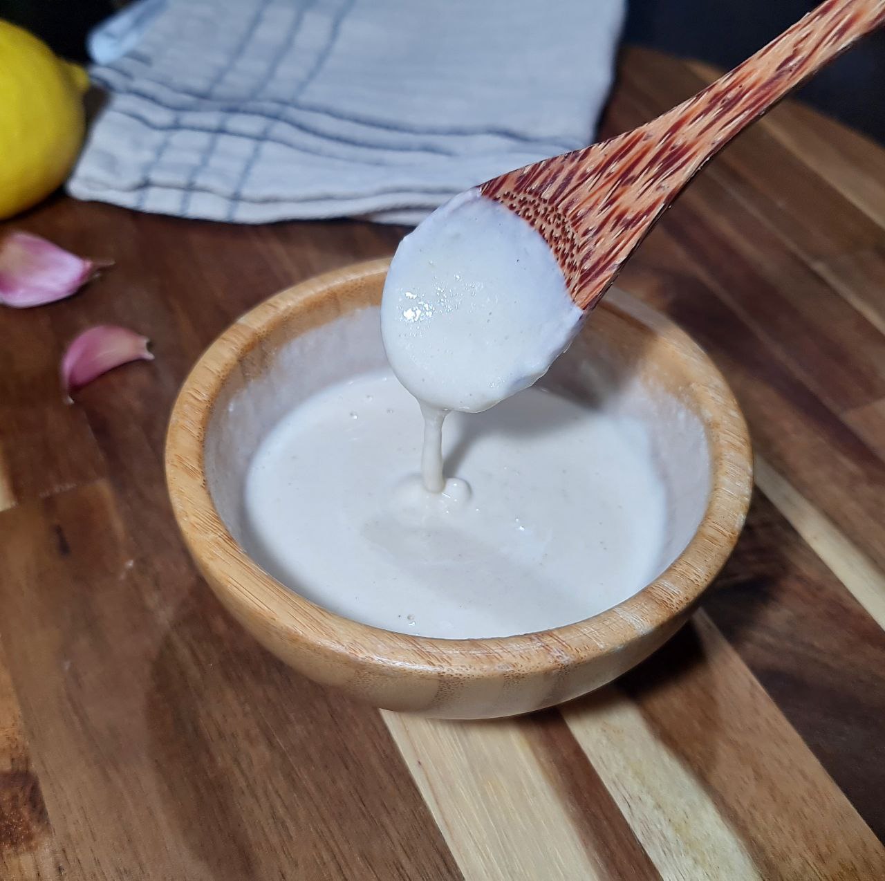 Spoon drizzling creamy tahini dressing into a wooden bowl. Creamy tahini dressing with a smooth texture being poured into a bowl.