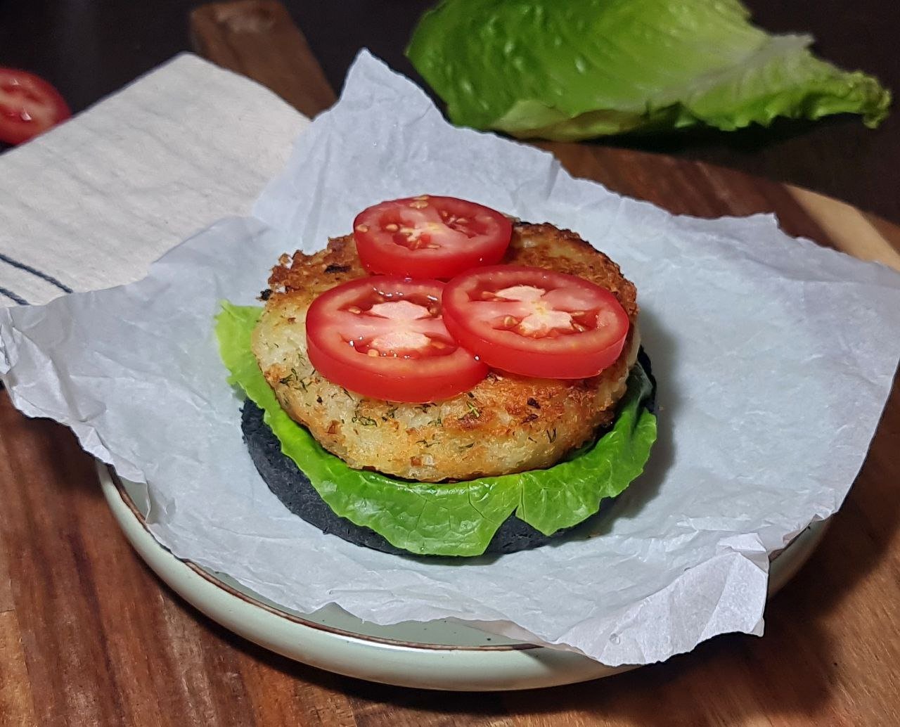 Partially assembled vegan potato burger with lettuce, potato patty, and tomato slices on a charcoal bun. Assembling the vegan potato burger with lettuce, a crispy potato patty, and fresh tomato slices.