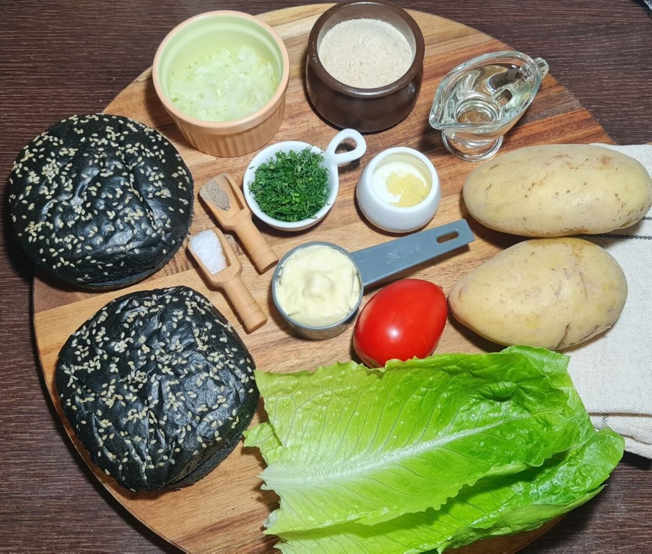 Ingredients for vegan potato burger, including potatoes, charcoal buns, lettuce, tomato, and seasonings. Fresh ingredients for making a vegan potato burger, displayed on a wooden board.