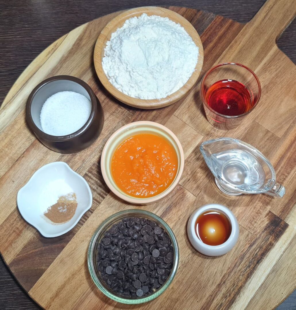 Ingredients for vegan pumpkin chocolate chip cookies displayed on a wooden board. Ingredients for vegan pumpkin chocolate chip cookies, including flour, pumpkin puree, chocolate chips, and spices.