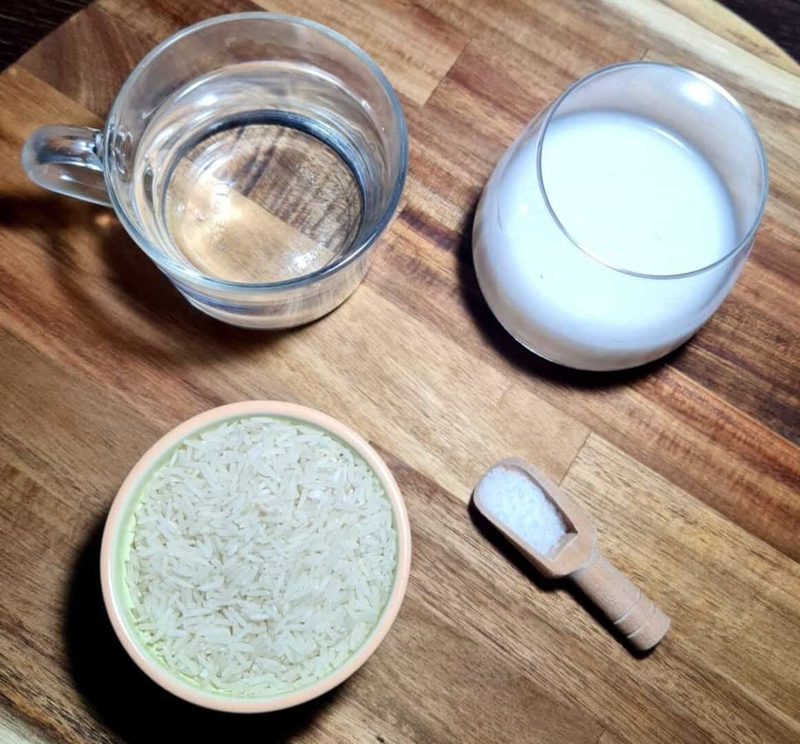 Clear glass mug with water, a round glass with milk, a small bowl with uncooked white rice, and a small wooden scoop with salt on a wooden surface.   