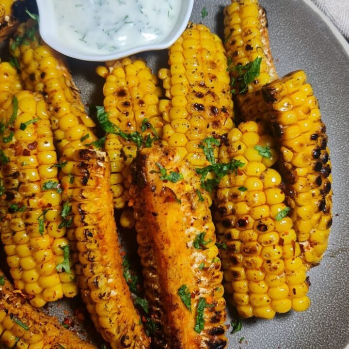 A plate of grilled corn ribs seasoned with spices and garnished with chopped herbs, served with a bowl of white dipping sauce. Grilled corn ribs seasoned with spices and garnished with fresh herbs, perfect for a flavorful snack.