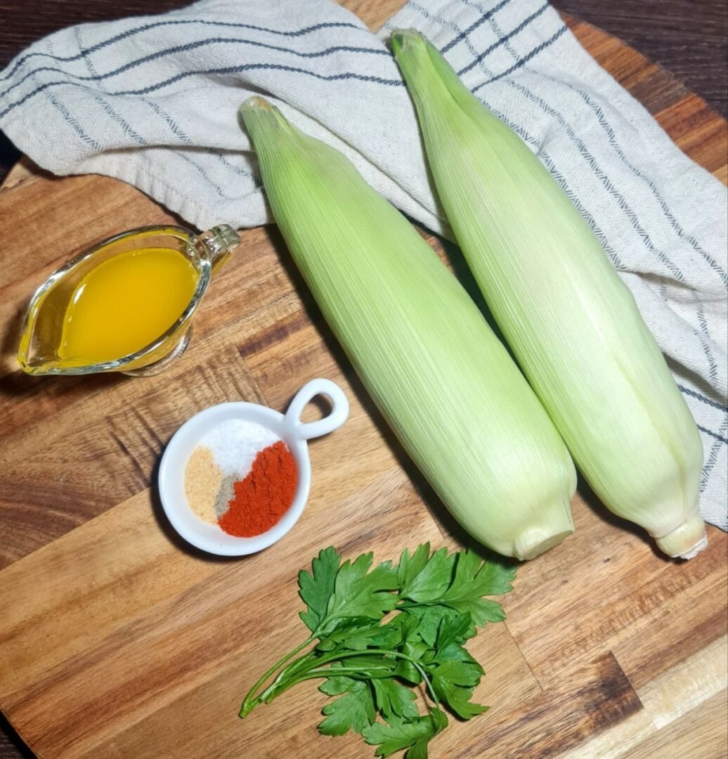  Wooden cutting board with corn in husks, oil, spices, and fresh parsley. Fresh ingredients for making delicious corn ribs.