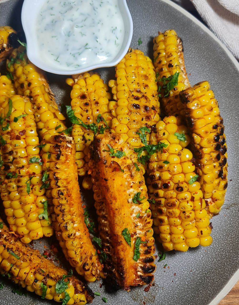 A plate of grilled corn ribs seasoned with spices and garnished with chopped herbs, served with a bowl of white dipping sauce. Grilled corn ribs seasoned with spices and garnished with fresh herbs, perfect for a flavorful snack.