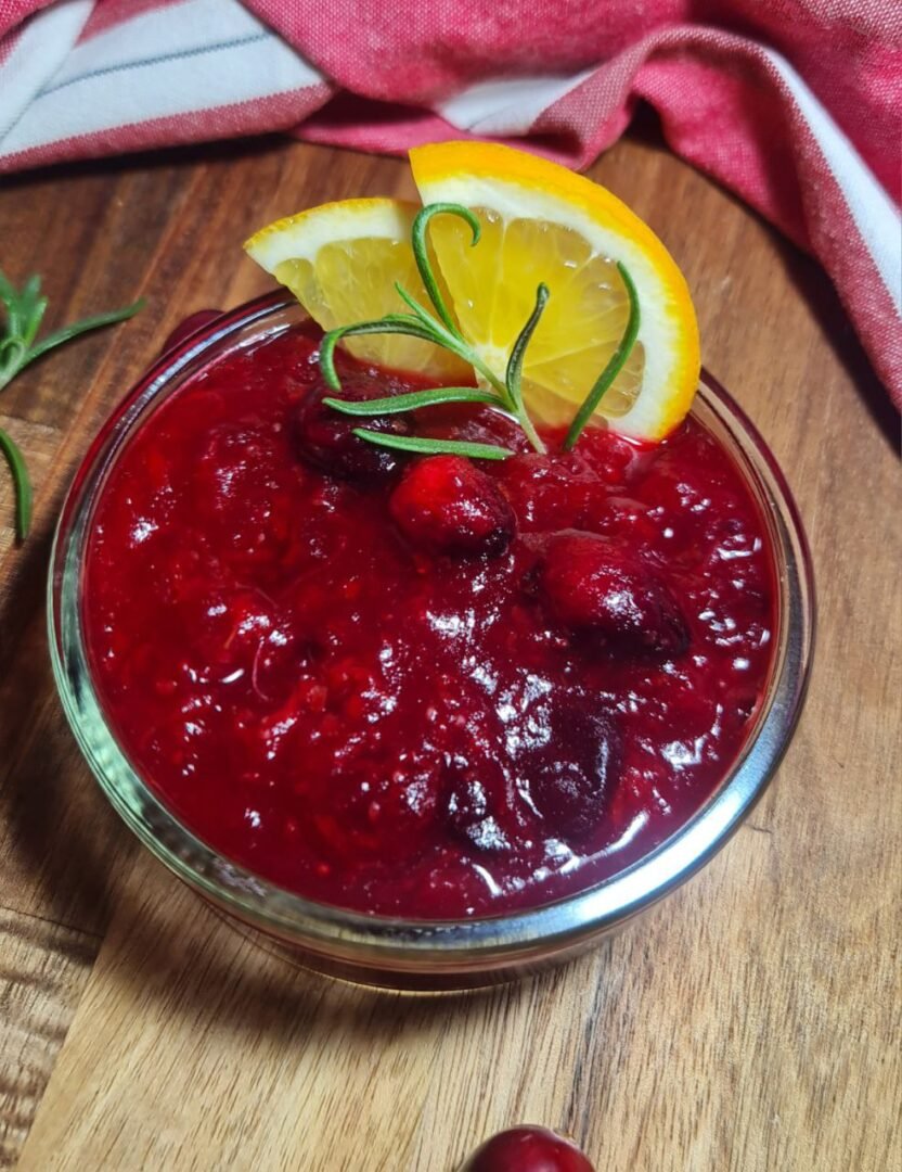 Bowl of homemade cranberry sauce garnished with orange slices and rosemary, surrounded by fresh cranberries on a rustic wooden surface.  Homemade cranberry sauce garnished with fresh orange slices and rosemary, perfect for holiday meals.