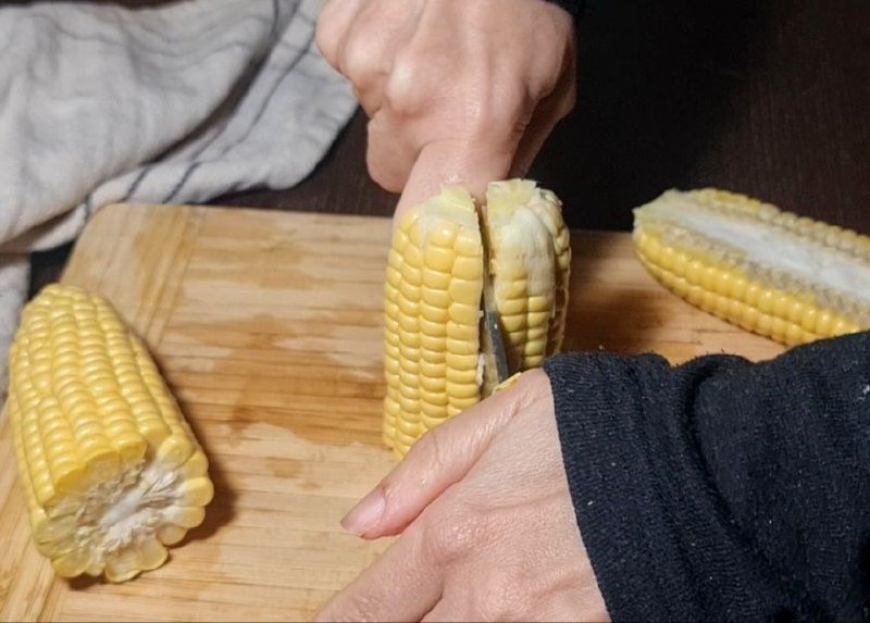 Hands cutting corn on the cob into smaller pieces on a wooden cutting board. Cutting corn on the cob into smaller pieces to prepare corn ribs.