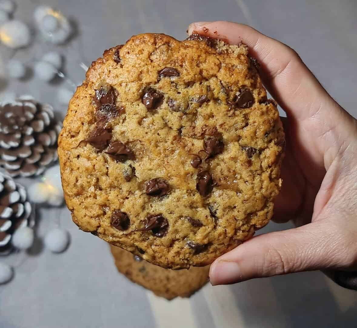 A hand holding a large vegan gingerbread chocolate chip cookie with a background of pinecones and festive decorations.