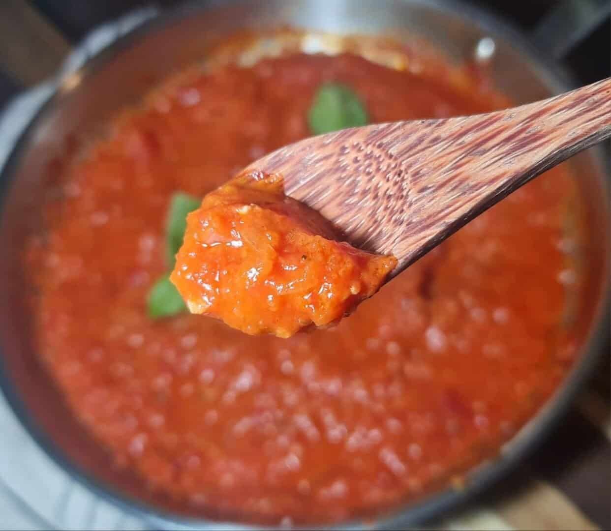 A close-up of a wooden spoon holding a scoop of homemade marinara sauce with a pot of sauce in the background.